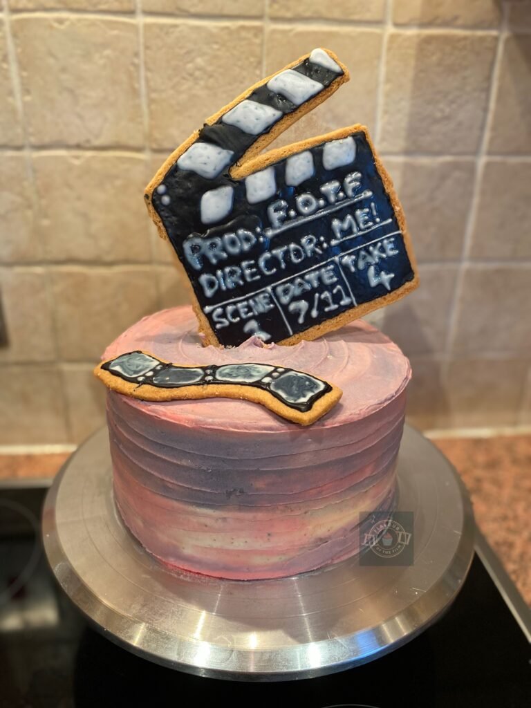 All images are of a three layer pink sponge cake with pale pink, lilac and white buttercream. The buttercream is applied on the cake to mimic a summer evening sunset. The cake is topped with a film clapperboard and a row of film cells that are both made out of shortbread biscuit. The biscuit toppers are decorated with royal icing that has been piped and dried. The royal icing reads as follows:
Prod: F.O.T.F.
Director: Me!
Scene: 1
Date: 7/11
Take: 4