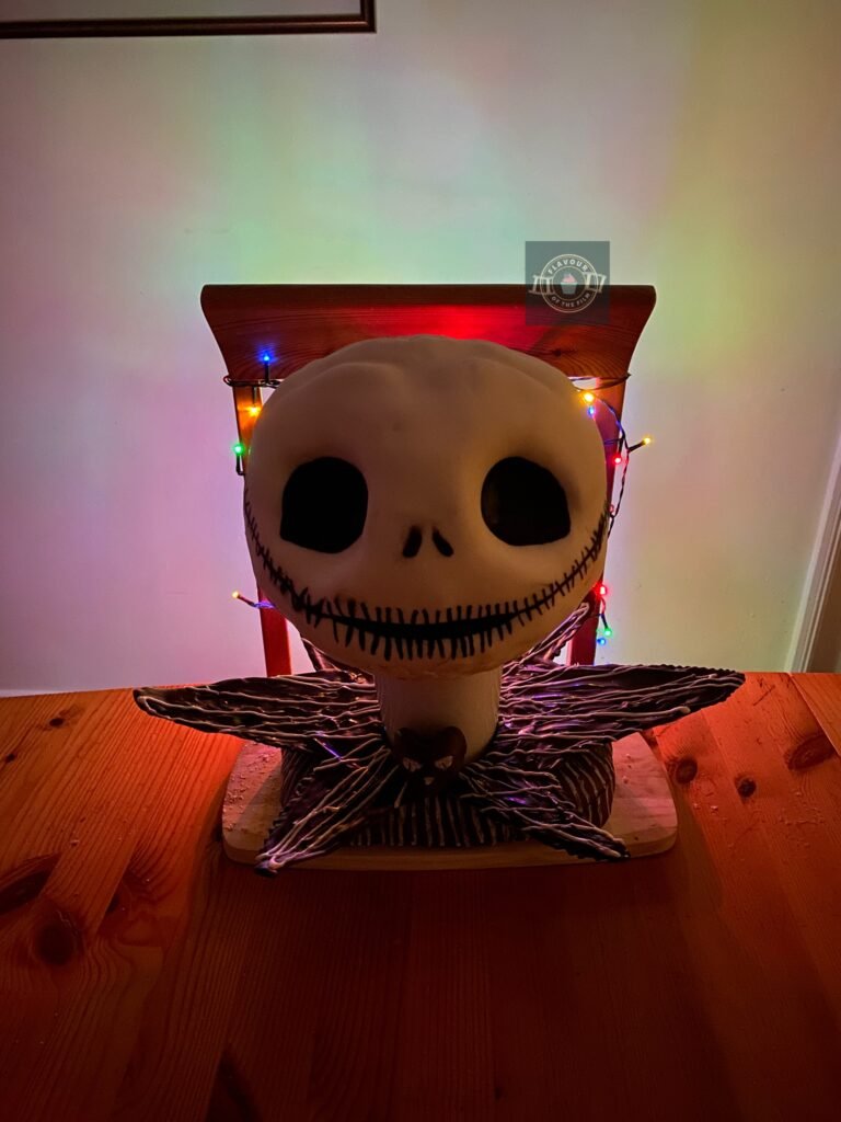All images are of a cake shaped and decorated with white fondant and edible paint like Jack Skellington's head, set on a white cup; a tempered chocolate collar, and spare cake in fondant covered block form. The cake bust is lit from behind by multi-coloured Christmas lights.