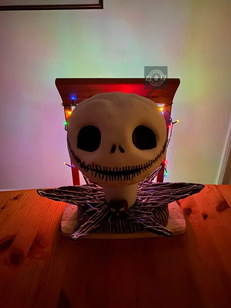 All images are of a cake shaped and decorated with white fondant and edible paint like Jack Skellington's head, set on a white cup; a tempered chocolate collar, and spare cake in fondant covered block form. The cake bust is lit from behind by multi-coloured Christmas lights.