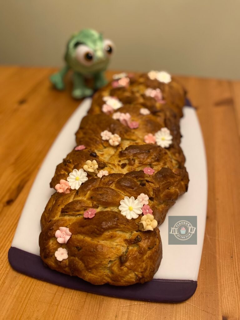 A golden braided brioche with candy flower decorations sits on a board with a Pascal plushie next to it. This bread celebrates Disney's Tangled.