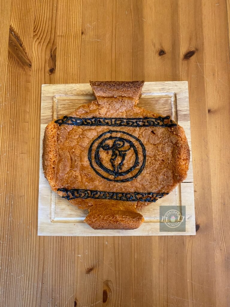All images are of a pale orange Greek vase shaped almond sponge which is decorated with ancient Greek patterning on the top and bottom edges of the vase's main body, as well as Hercules's lightning bolt medallion in the centre. All decoration is piped in black royal icing. The vase is in two layers that are sandwiched together with honey and lemon curd. This vase Bakewell celebrates Disney's Hercules.