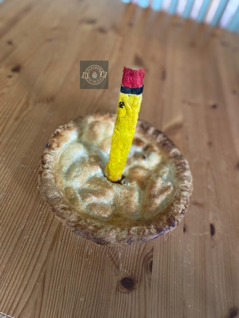 All images are of a golden double crust pastry apple and blackberry pie with a pastry HB pencil sticking out of its lid that has been painted in yellow with a pink end. This pie celebrates the John Wick series.