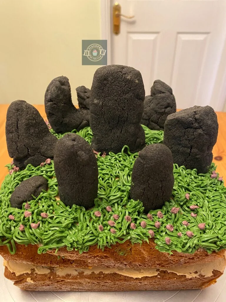 All images are of a two layer coffee and walnut traybake cake sandwiched together by coffee buttercream and decorated with piped grass and purple flowers on the top of the cake and grey shortbread shaped and arranged in the form of Craigh na Dun, Scotland.