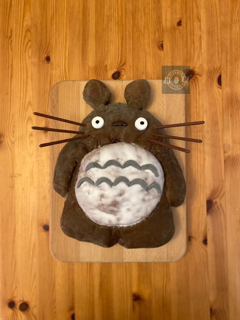 All images are of a bread loaf shaped like the character Totoro from the Anime film, My Neighbor Totoro. The bread is topped with royal icing for his belly and fondant facial features. Plus, Pocky chocolate sticks to make his whiskers.