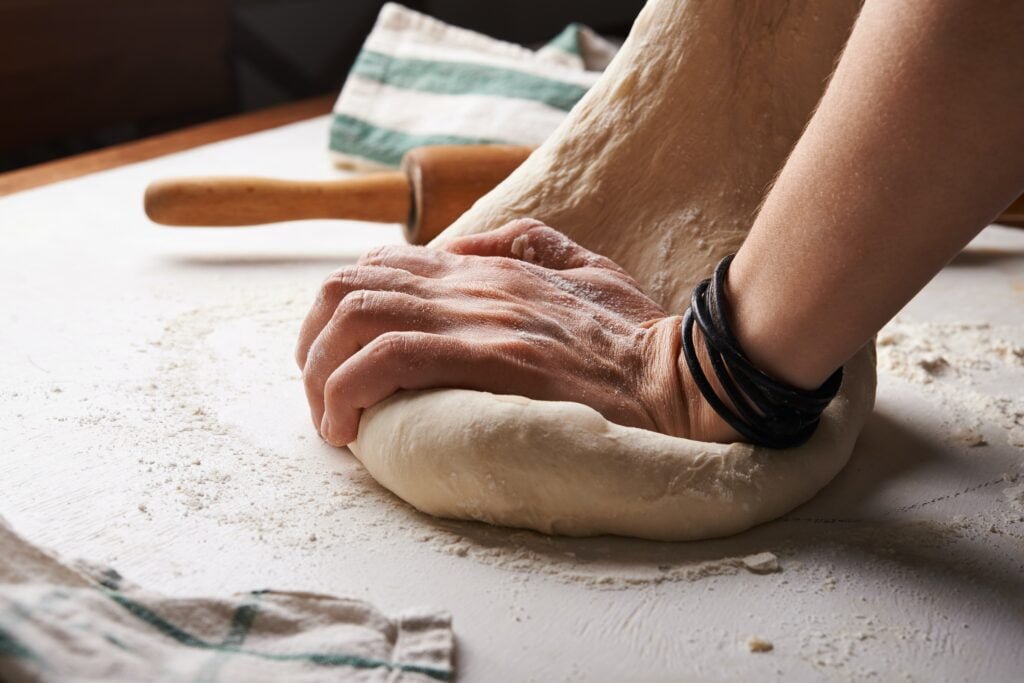 A pair of hands are stretching and kneading some bread dough. There is a green and white striped tea towel and a rolling pin in the image, too. This image goes with the window pane test section of my top 10 baking tips.