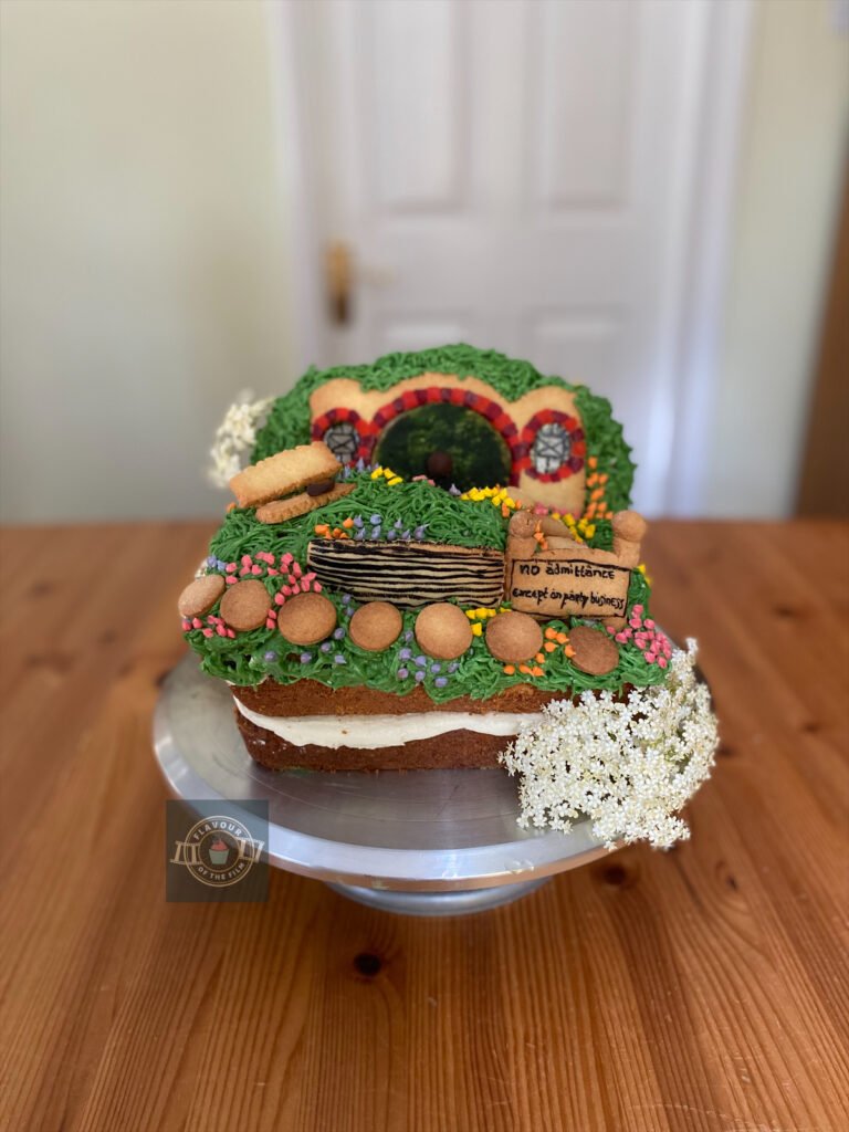 All images are of a square lemon and elderflower landscape cake that is decorated with buttercream, extra cake and biscuit decorations all in the shape and look of the hobbit hole known as Bag End, celebrating The Hobbit films. The cake is finished with the flowers from an elderflower tree.