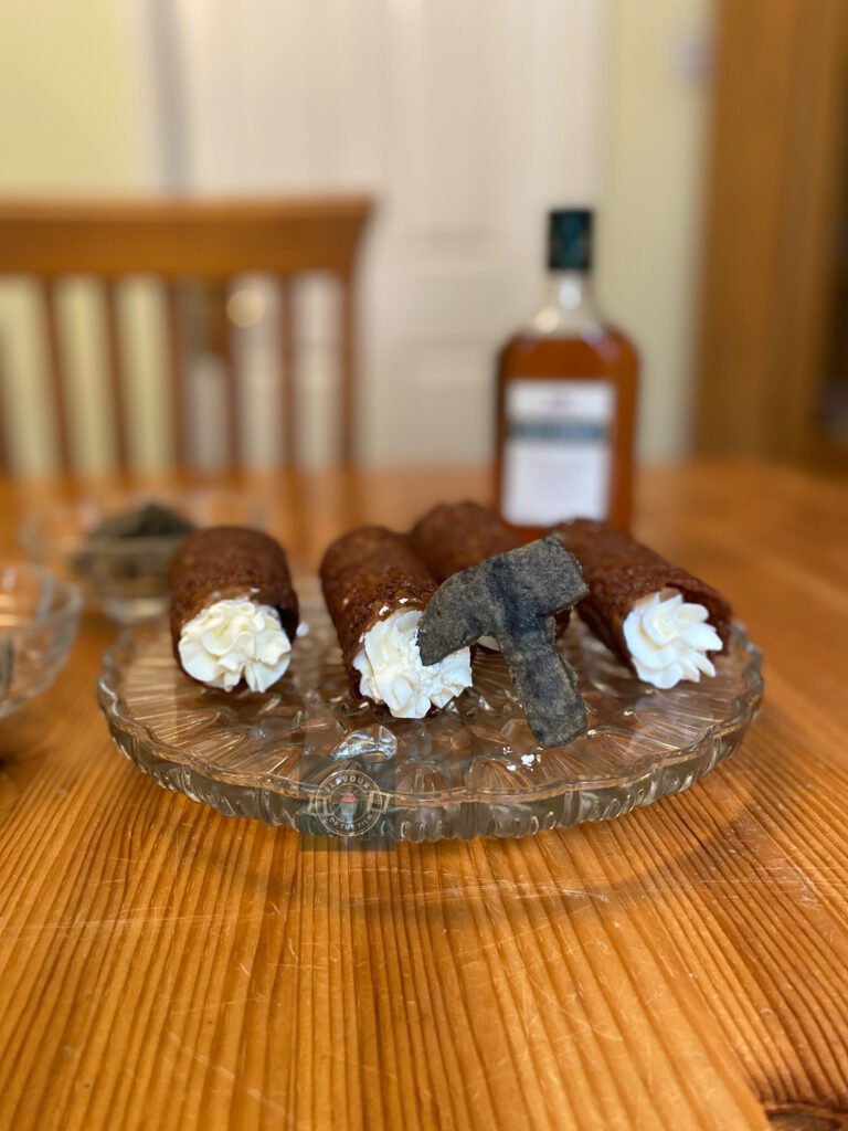 All images are of four dark brandy snaps filled with piped whipped cream and pick axe shaped grey shortbread. The brandy snaps are on a glass serving plate and the shortbread pick axes are in glass bowls. There is a bottle of brandy in the background.