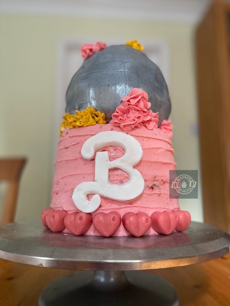 All images are of a two tiered Barbie themed cake. The base cake is covered in baby pink buttercream with pink chocolate hearts forming a collar around the bottom edge of the cake. There is the iconic Barbie 'B' shaped in white fondant icing that is sitting on the front of the base cake. The top tier is a disco ball made from pink champagne cake and decorated with silver fondant icing, and pink and yellow buttercream rosettes on the top and around the bottom of the disco ball. The entire two tier cake is glistening in the natural light thanks to the edible spray glitter.