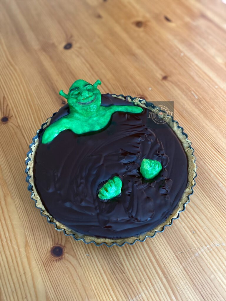 All images are of a salted caramel and chocolate tart encased in a biscuit case. Shrek is sitting in the chocolate of the tart. Shrek is made entirely out of white modelling chocolate, and is airbrushed green with hand painted detailing. The only visible parts of Shrek are his head, shoulders, upper arms and feet.
