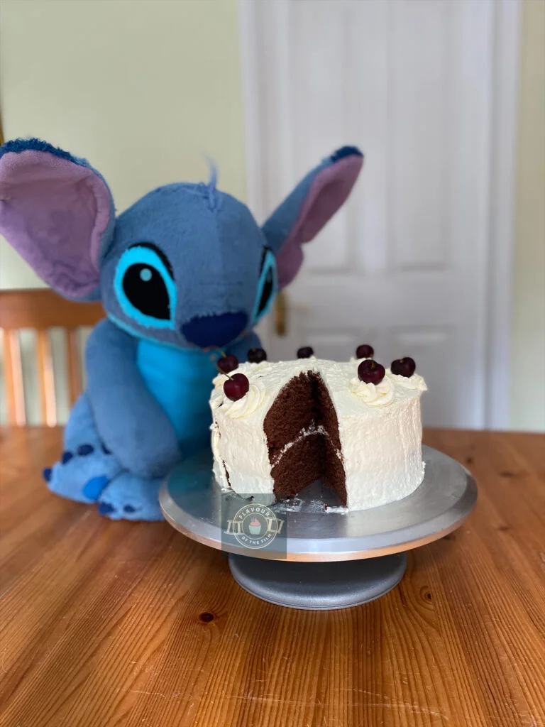 All images are of a two layer chocolate cake which is covered in a white Swiss meringue buttercream. The cake is topped with eight swirls of buttercream and a fresh whole cherry on each swirl. A giant plushie version of Stitch is is in the background, looking at the cake.