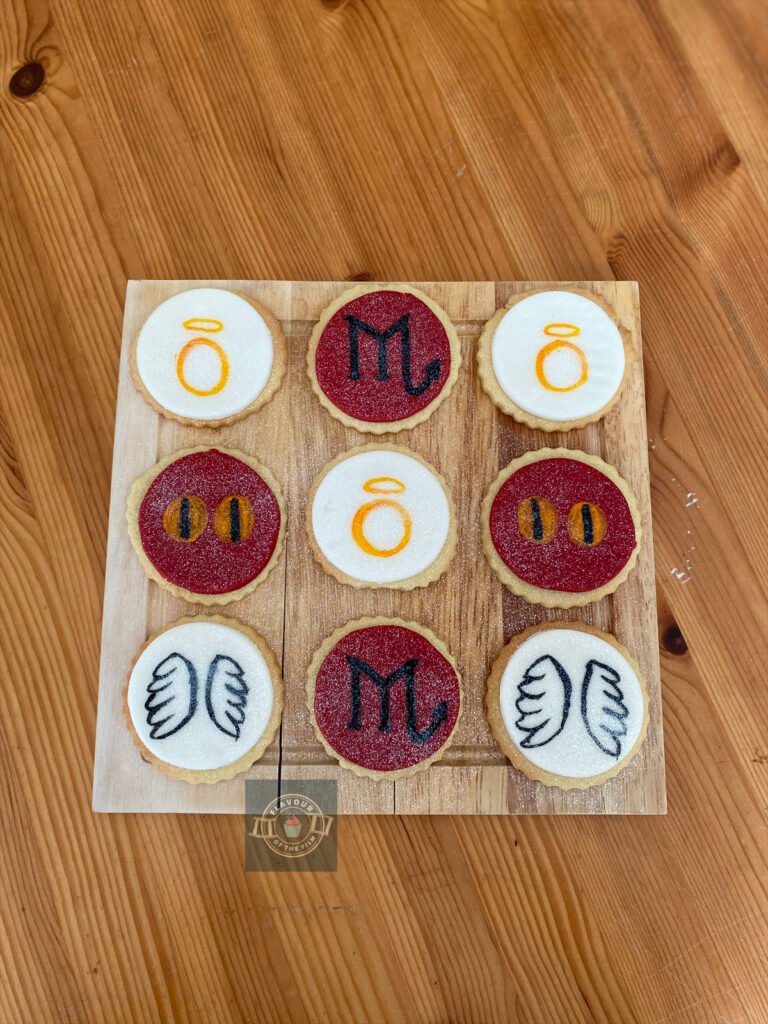 All images are of a wooden board holding 9 golden sugar cookies topped with alternating white and red fondant. The red fondant is hand painted with Crowley's golden devil eyes and the 'M' from the Good Omens title font. The white fondant is hand painted with the 'O' from the Good Omens font and a set of angel wings. All cookies are finished with edible glitter.