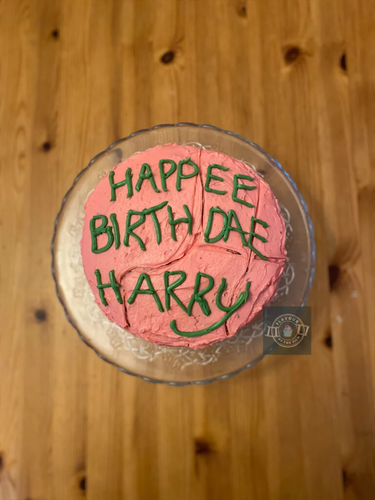 All images are of a two layer chocolate fudge cake that is covered in baby pink buttercream and topped with the iconic misspelled birthday with to Harry from Hagrid, piped in green buttercream handwriting. The cake is on a glass cake stand which the author placed in a white box in some images in order to recreate the near exact visuals from the film.