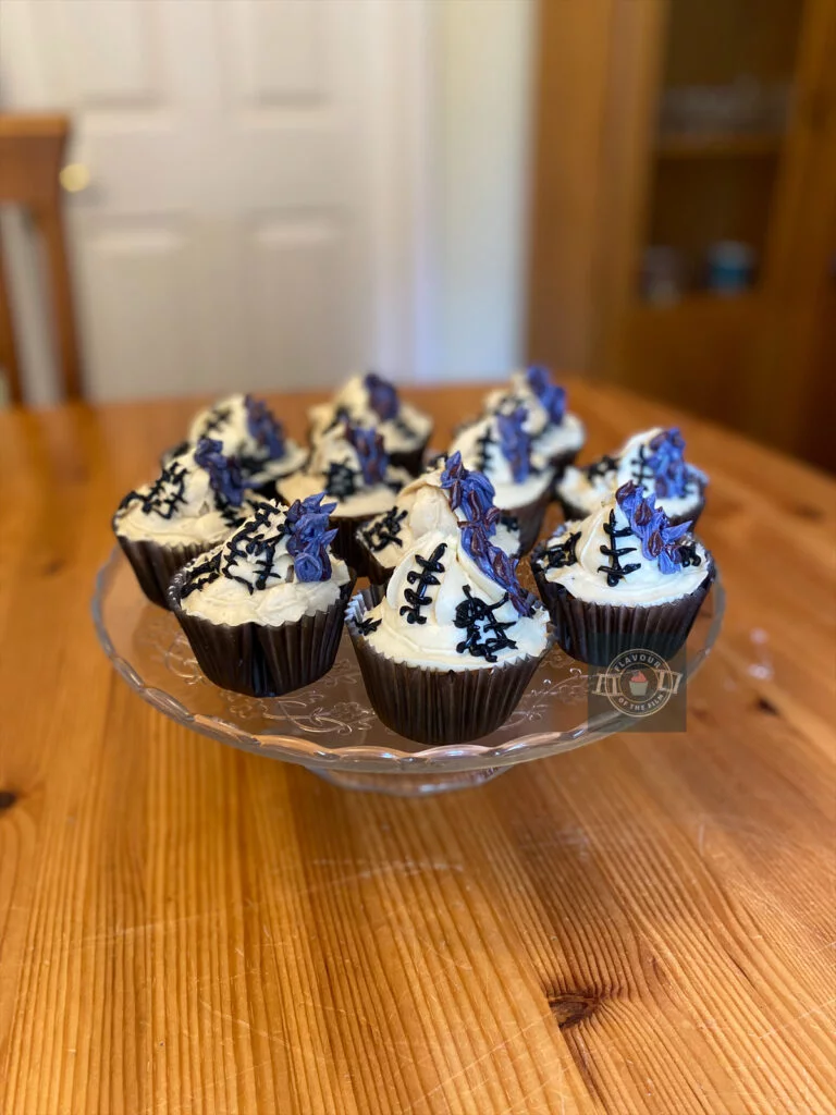 All images are of cupcakes in black cupcake cases with pale buttercream that has been piped in the shape of a witch hat on each cake. The decoration is finished with purple buttercream features and black buttercream stitching. The cupcakes are filled with red butterscotch sauce.