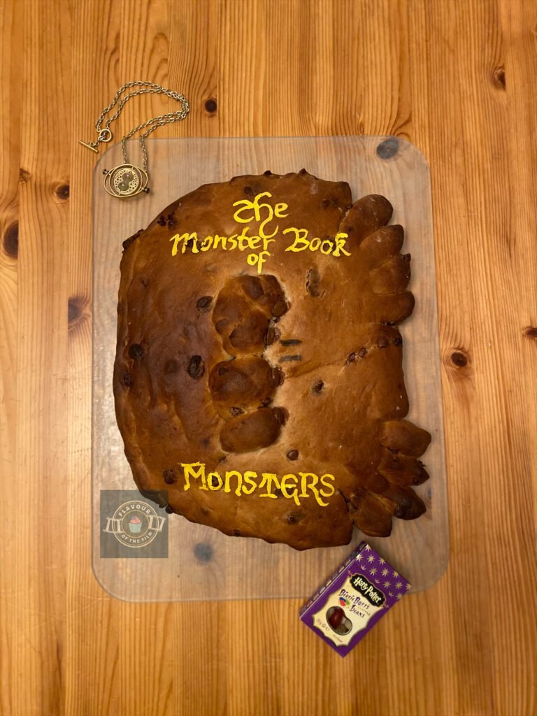 All images are of a golden loaf of bread shaped to look like the Monster Book of Monsters. The eyes are chocolate chips, which are also mixed into the bread loaf itself. The decoration is hand painted in gold and reads "The Monster Book Of Monsters" above and below the book's eyes. The loaf rests on a transparent board and is surrounded by a copy of the Marauders map; Bertie Botts every flavour beans, and Hermione's time turner. This bread loaf celebrated Harry Potter and the Philosopher's Stone.