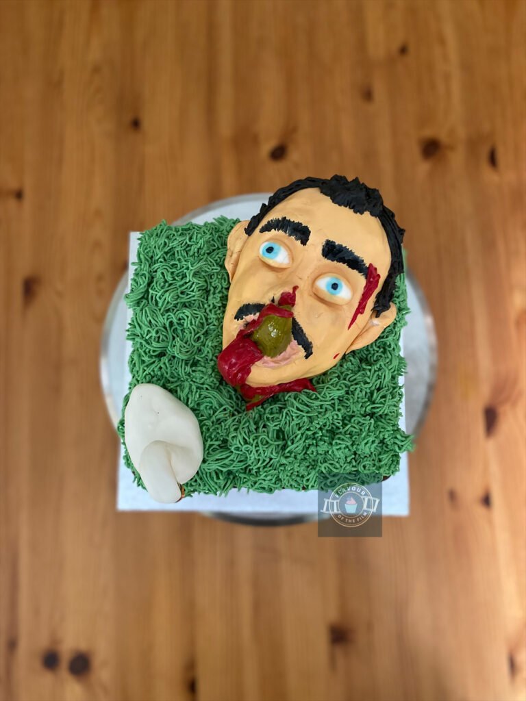 All images are of a two layer square landscape cake, with cherry jam and fresh whipped cream oozing out from between the layers. The cake is topped with green grass made from fresh whipped cream. To decorate, the cake has been topped with a fondant swan and a fondant head that has been painted to resemble Timothy Dalton with a fondant steeple through his mouth. This landscape cake celebrated Hot Fuzz.