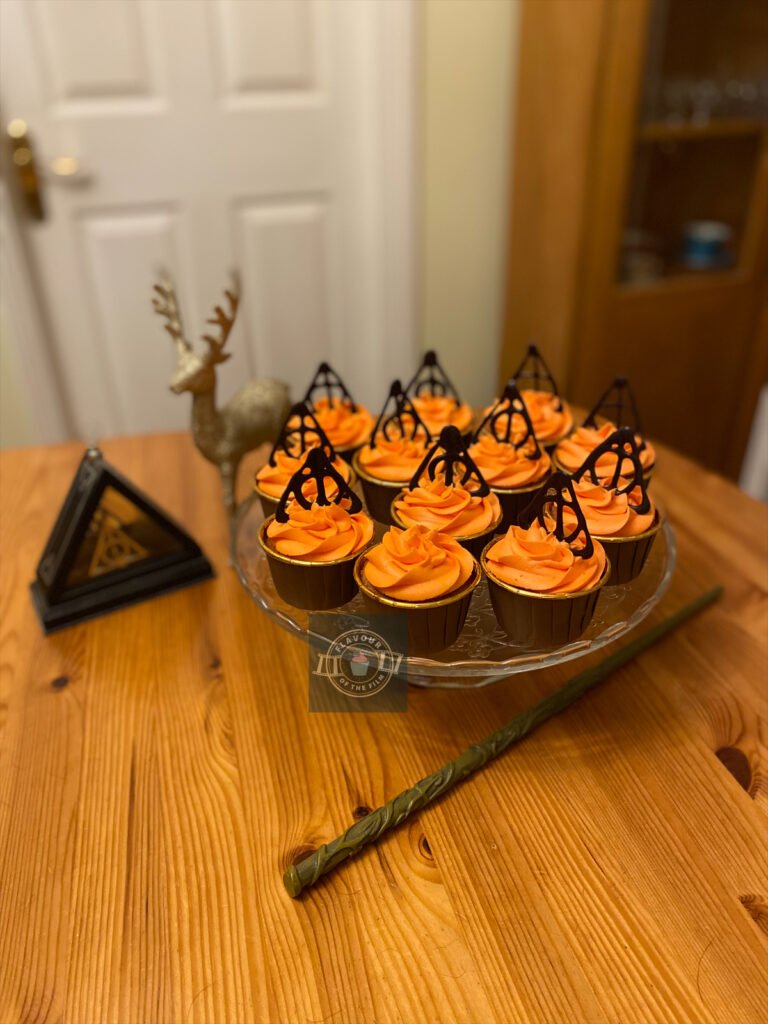 All images are of 12 cupcakes in gold and brown cupcake cases, topped with vibrant orange swirls of buttercream and tempered dark chocolate Deathly Hallows symbols. The cupcakes are presented on a glass cake stand. Hermione's wand, a Deathly Hallows necklace in a triangle glass case and a gold glittery stag are decorating the background.