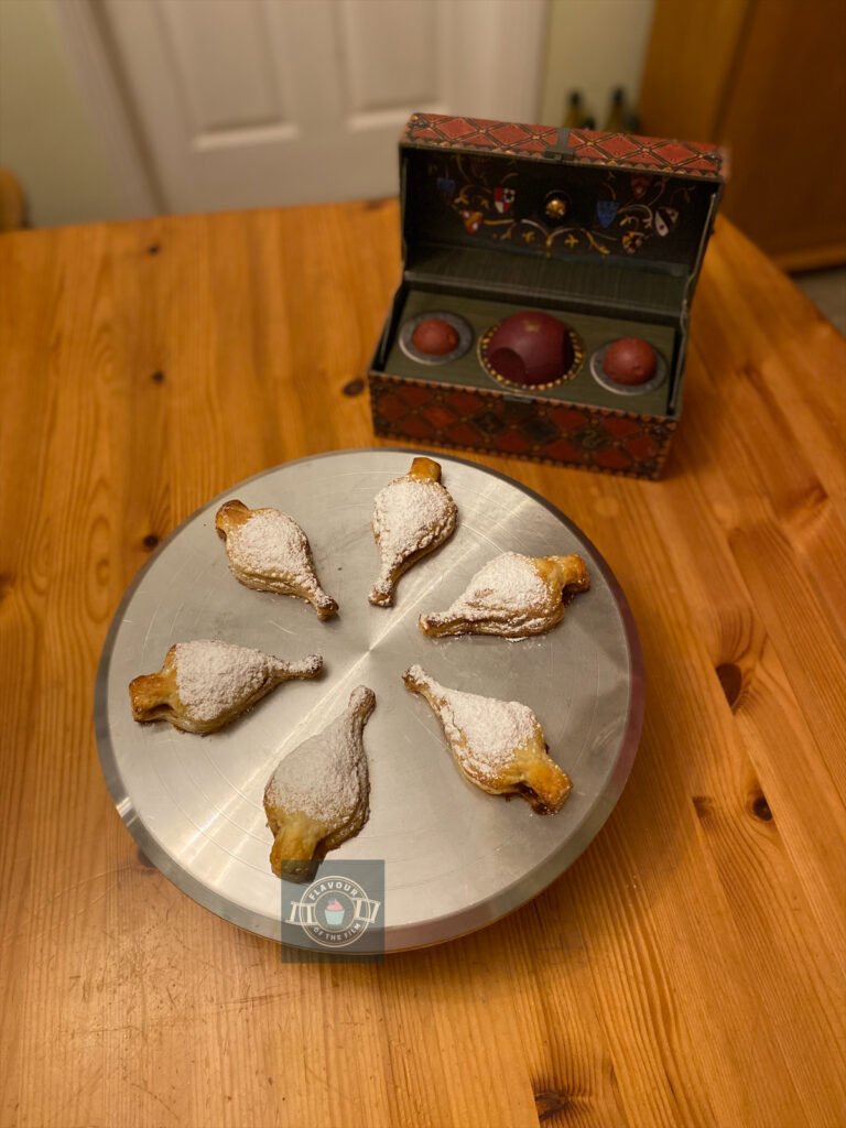 All images are of puff pastry turnovers in the shape of liquid luck bottles. They are each dusted with icing sugar and are presented on silver metal cake stand. There is a smaller version of the quidditch equipment case in the background. These pineapple turnovers celebrate Harry Potter and the Half-Blood Prince.