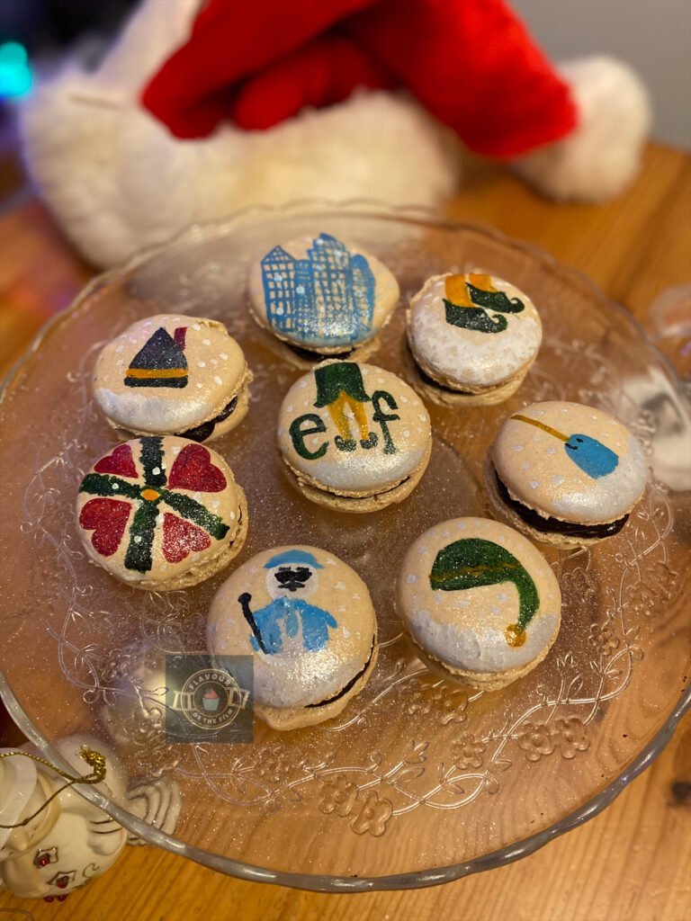 All images are of white macarons with chocolate filling and are hand painted with decorations related to Elf. The macarons give a similar effect to snow globes. The painted imagery include Buddy's elf hat, Buddy's elf feet on snow, Jovie's elf hat, the narwhal in the ice, the heart pattern on Santa's suspenders, the Empire State Building, and Leon the snowman. The macarons are presented on a patterned glass cake stand and are surrounded by a Santa hat, Santa figurines, some red and gold Christmas baubles and a snowman figurine. There are multi-coloured Christmas lights in the background, too.