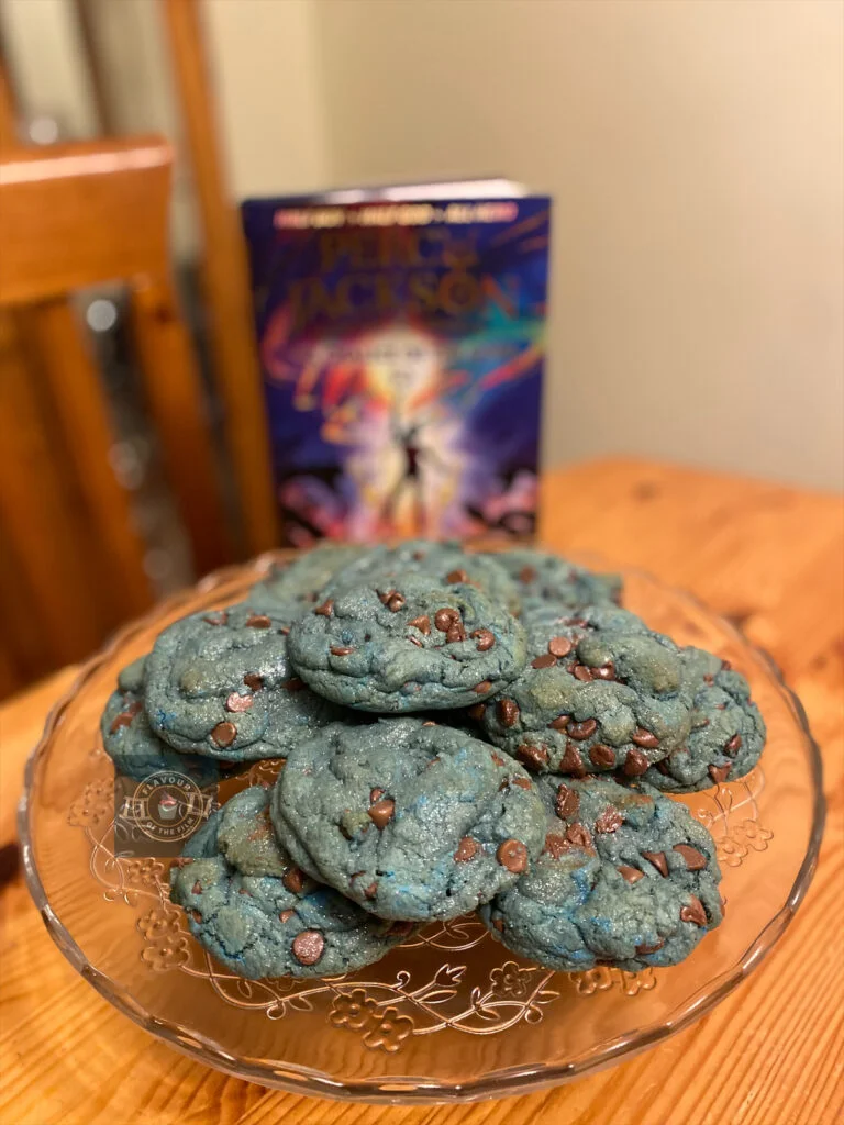 All images are of a batch of 12 seafoam blue chocolate chip cookies. The chocolate chips are milk chocolate, and the base of the cookies are thick NYC-style. The cookies are presented on a glass cake stand, with the latest book addition to the Percy Jackson series.