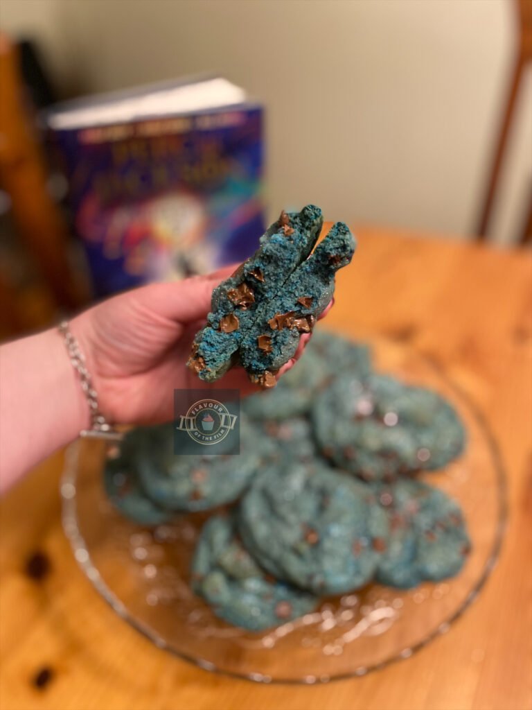 A blue chocolate chip NYC style cookie in halves and held in author's hand, with the rest of the cookies in view. These cookies celebrate Percy Jackson and the Olympians.