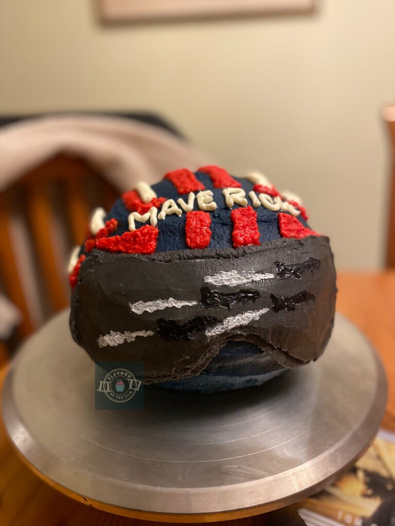 All images are of a spherical two layer cake with navy, white and red buttercream piped on to resemble Maverick's helmet in Top Gun: Maverick. The visor is hand painted to give the impression that four fighter planes are flying by. The cake is presented on a silver metal cake stand, with a pilot's jacket and a DVD copy of the film in the background. "Maverick" is piped in buttercream in white on the helmet cake.