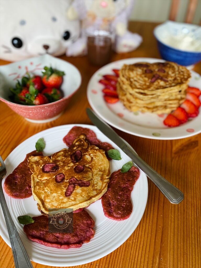 All images are of a single pancake on one plate, surrounded by eggplant/aubergine shaped pancakes. There is a stack of the same shaped cute pancakes on a white plate with pastel polka dots slightly behind the singular pancake. The stack is surrounded by sliced strawberry pieces. There is also a pink cat shaped bowl containing fresh strawberries and a blue cat shaped bowl containing sweetened mascarpone and a whisk. A small pot of Nutella is also in the shot, as are a lilac bunny plush toy and a cute seal plush toy. The pancakes are in the shape of a grumpy cat face with pink features, celebrating Bee and Puppycat.