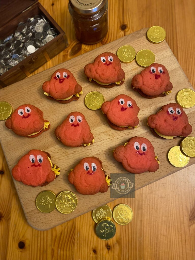 Individual marmalade biscuits with gold buttercream and marmalade fillings. Each biscuit is a slightly different shape and is made from a rich pink-coloured dough. The top of each biscuit is hand painted with cute eyes and a smiling mouth. The biscuits are placed neatly on a wooden board and are surrounded by pirate gold coins and a treasure chest of silver coins.