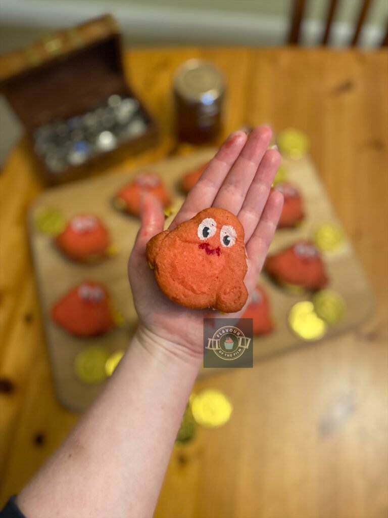 Author's hand holding one of the individual marmalade biscuits. The rest of the biscuits are in the background, with the pirate gold coins, a jar of marmalade and the treasure chest of silver coins.