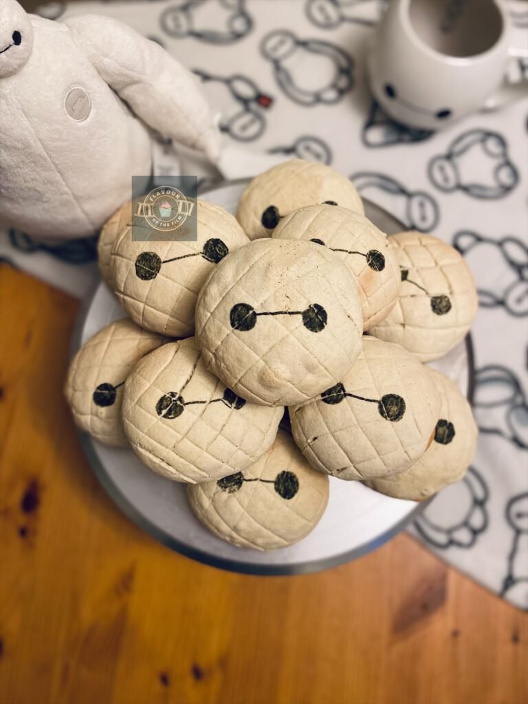 All images are of round sweet bread buns with a white melon like criss-cross pattern and the features of Big Hero 6's Baymax drawn on: his round eyes and connecting line between them. There are 12 buns in total, presented in a pile on a silver metal cake stand. In the background, there is a white Baymax print blanket covering half of the table beneath the buns; a large round Baymax mug, and a medium sized Baymax plushie.