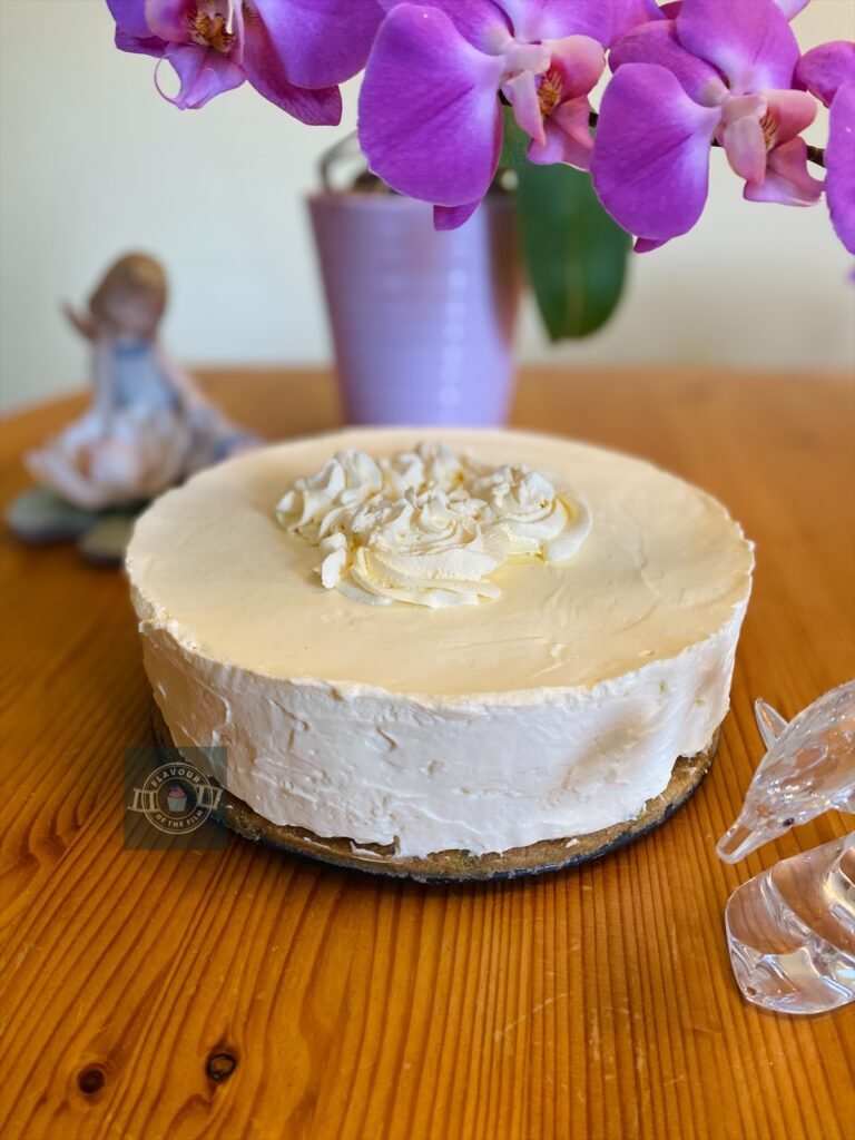 All images are of a pale cream coloured cheesecake on a brown biscuit base. The cheesecake is topped with four swirls of whipped cream that have been sprayed with edible gold glitter. Some images are of a thick slice of the cheesecake on a white plate with a silver fork. In the background, there is a lilac pot of purple orchids; a ceramic statue of a young girl sitting on a lily pad with a lotus flower, and a small crystal dolphin.