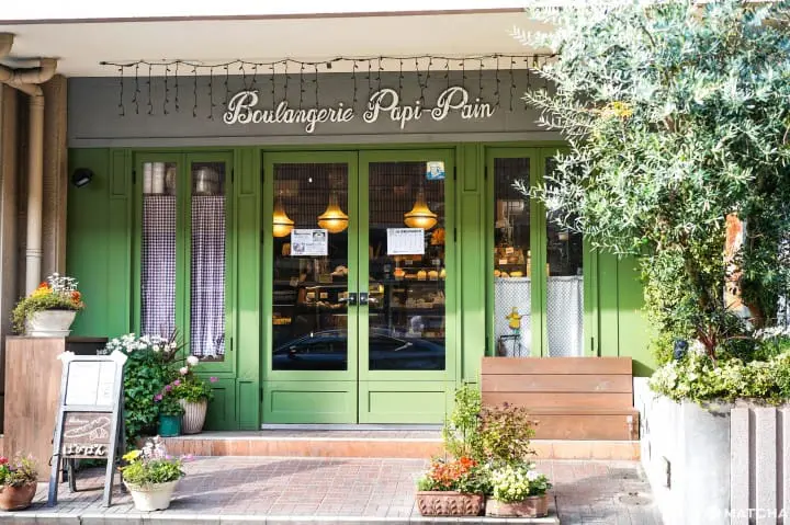 Exterior of Boulangerie Papi-Pan, Shizuoka. Green door frames around a double door entrance with large windows. Trees, a wooden bench and potted flowers sit all around the entrance. The bakery's sign is in script-style writing and sits above the double door entrance, with fairy lights hanging down in front of it.