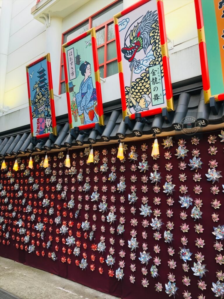 Multi-coloured pinwheels pinned on a wall by Asakusa Kagetsudo. Framed artwork of a dragon, a Geisha, and Senso-ji temple hang above the pinwheels.