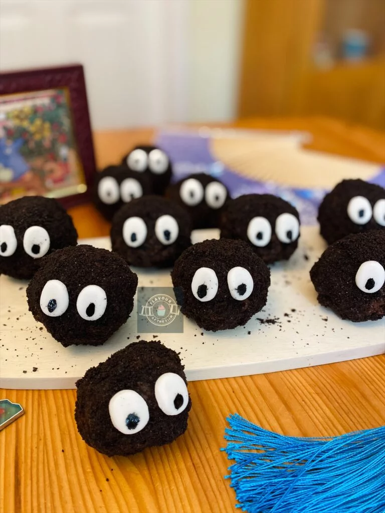 All images are of triple chocolate cookie dough balls covered in a dark Oreo cookie crumb, finished with two white oval eyes with black pupils. Some of the cookie dough bites are displayed on a rounded regtangular pale wooden plank, with the rest dotted around. Props include a stained glass window-esque framed puzzle of Totoro on a tree branch; a blue Japanese fan keyring, a lucky Japanese cat with blue tassle, and a purple Japanese fan with blue butterflies on.