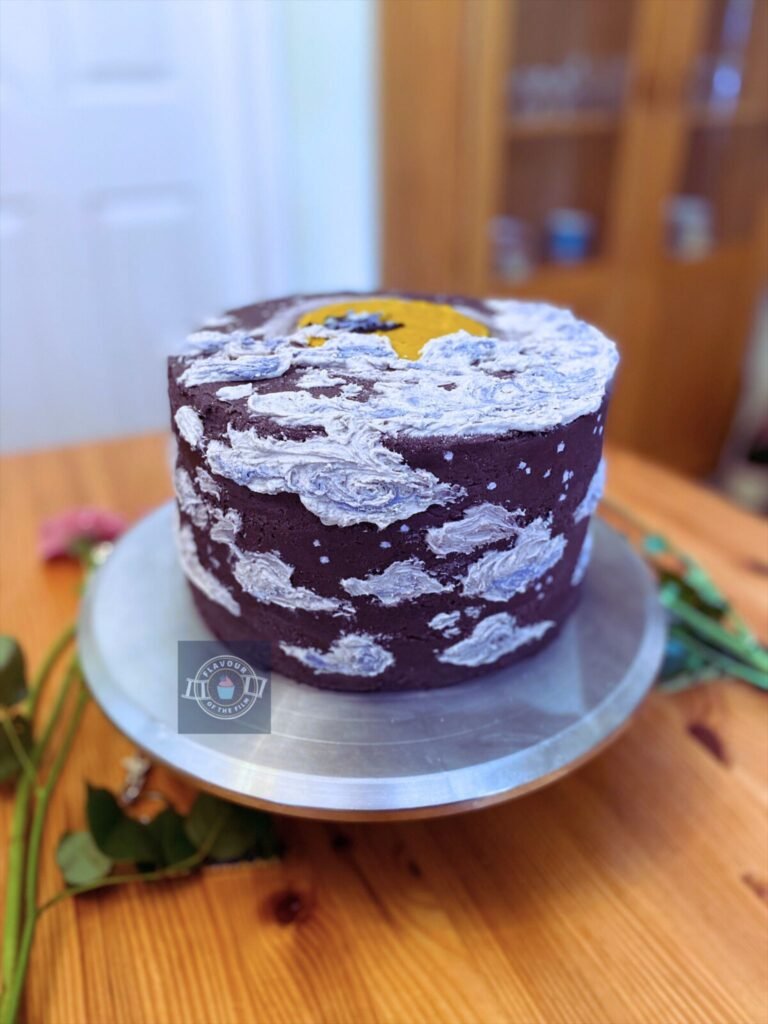 All images are of a purple three layer vanilla cake with cookies & cream flavouring. The cake is purple all over with white and blue whispy clouds painted on in buttercream; a yellow buttercream sun, and Aladdin & Jasmine silhouettes flying on the magic carpet into the sunset. The cake is displayed on a silver cake stand. Props include pink flowers, a white and pink rose, purple flowers, an Aladdin keyring, and a plushie of Genie in bee form.