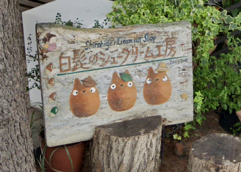 Rustic wooden sign outside Shiro Hige's Cream Puff Factory with hand painted Totoro shaped cream puffs on it. The sign is wedged inside a tree stump and is surrounded by growing greenery like ivy.