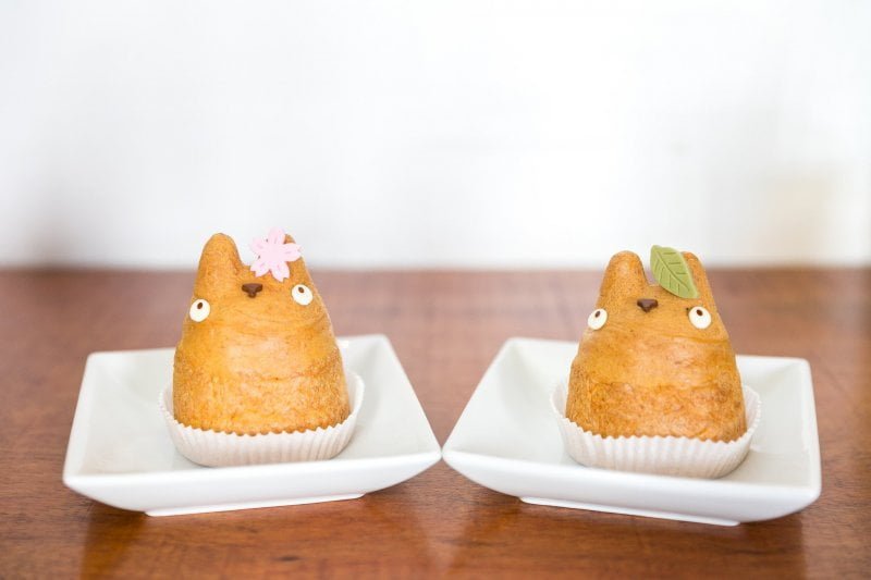 A pair of Totoro shaped cream puffs from Shiro Hige's Cream Puff Factory. One has a sugar paste cherry blossom tree flower on an ear, the other has a sugar paste green leaf in the same place.