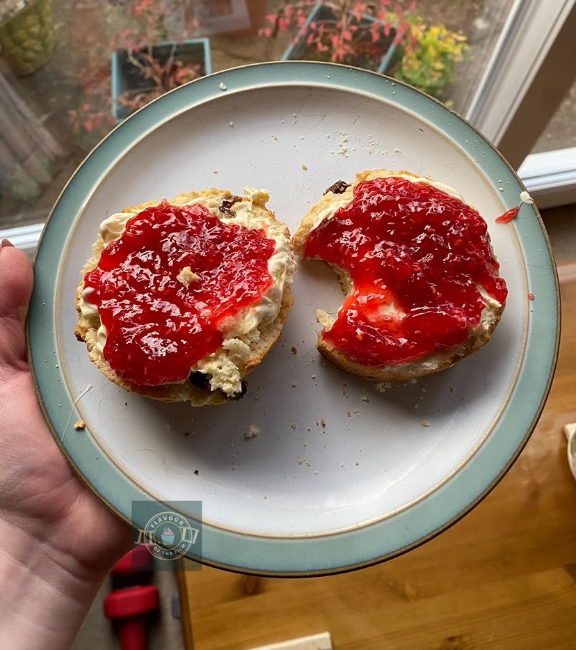 My homemade scone on a plate topped with clotted cream and jam. One half of the scone had a bite taken out of it and looks like a crescent moon.