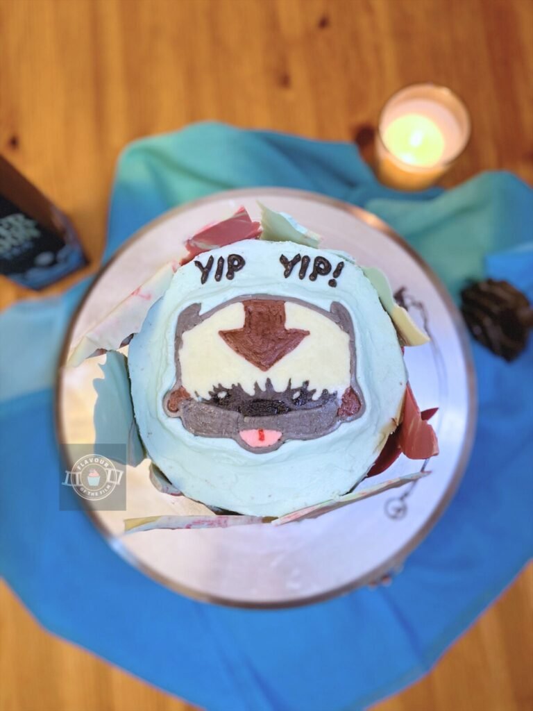 All images are of a light blue bento (6 inch, two layer) round cake with multi-colour marbled chocolate shards donning the outside and Appa's face made out of buttercream on the top of the cake. "YIP YIP" is piped in black buttercream above Appa's face. The cake is displayed on a silver metal cake stand. Props include a blue ombre scarf, a wave pendant necklace, a lit candle, "Silver Moon" tea, a toy boulder, and a bracelet with beads representing all of the chakras.