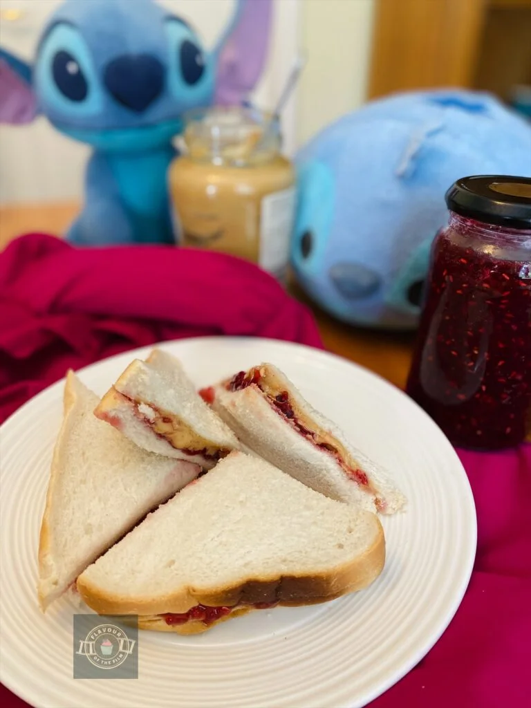 Four triangle shaped peanut butter and jam sandwiches displayed on a small plate in front of two Stitch plushies, a jar of peanut butter and a jar of my homemade jam.