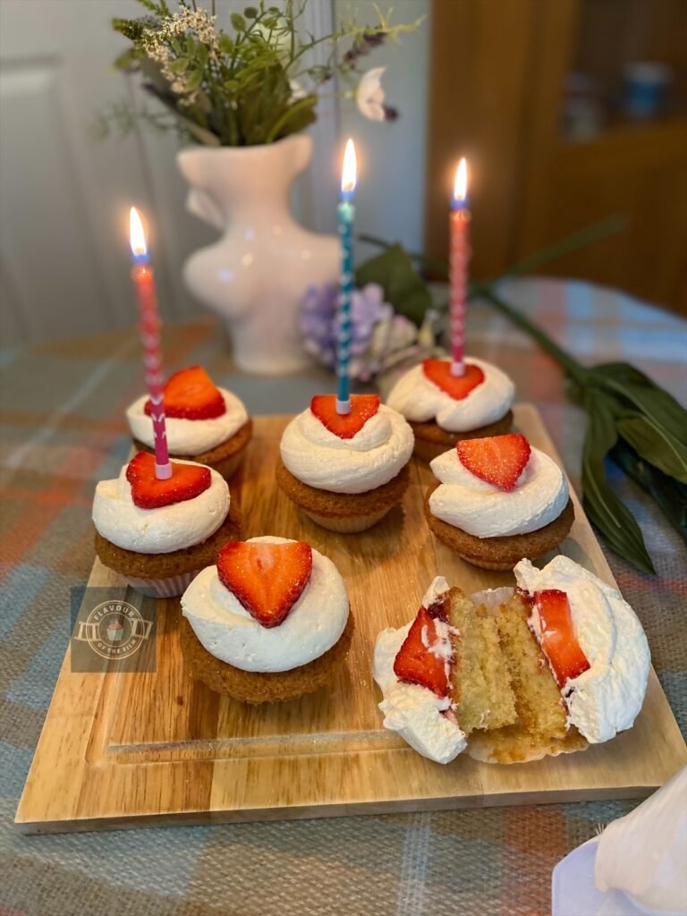 Vanilla cupcakes with fresh cream and a fresh strawberry slice on top, displayed on a wooden board with wildflowers around.