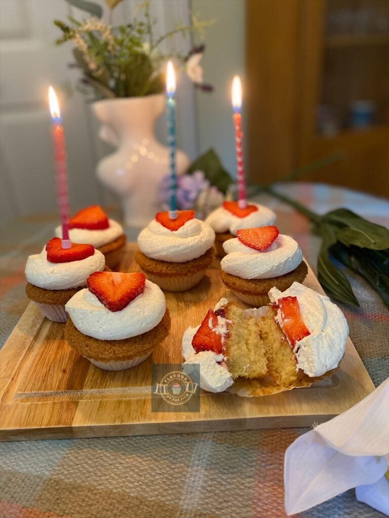 Vanilla cupcakes with fresh cream and a fresh strawberry slice on top, displayed on a wooden board with wildflowers around.