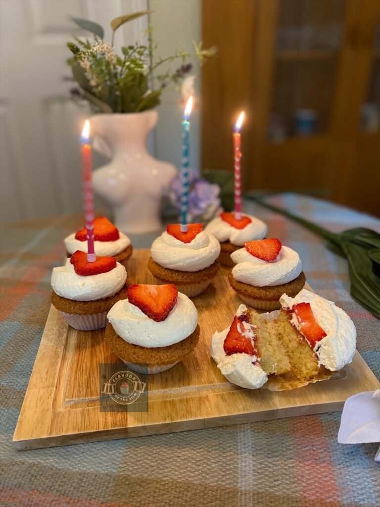 Vanilla cupcakes with fresh cream and a fresh strawberry slice on top, displayed on a wooden board with wildflowers around.