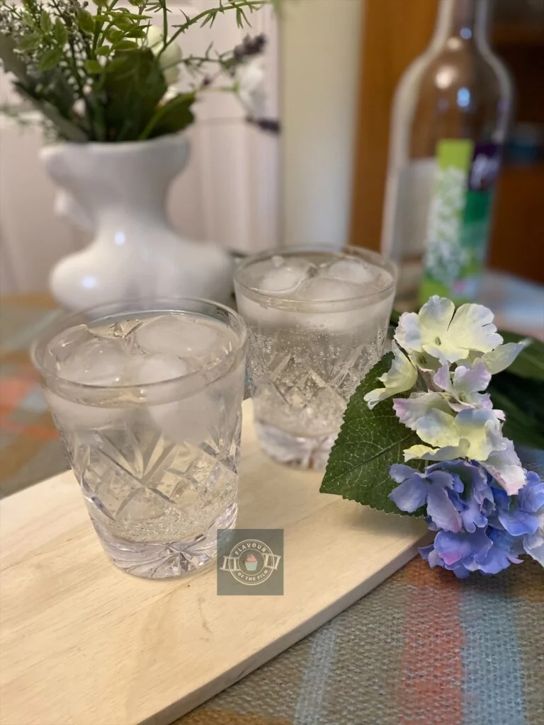 Two crystal glasses of elderflower presse with ice, displayed on a wooden board with wildflowers around.