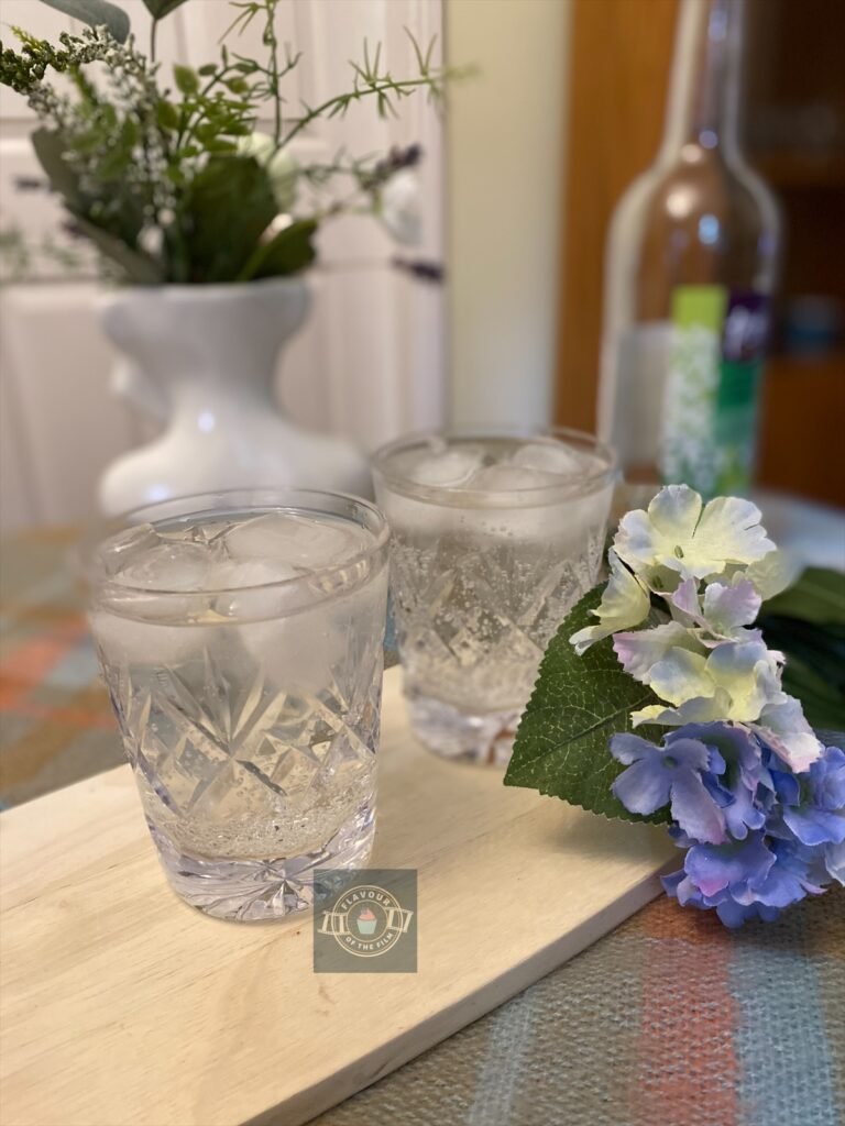 Two crystal glasses of elderflower presse with ice, displayed on a wooden board with wildflowers around.