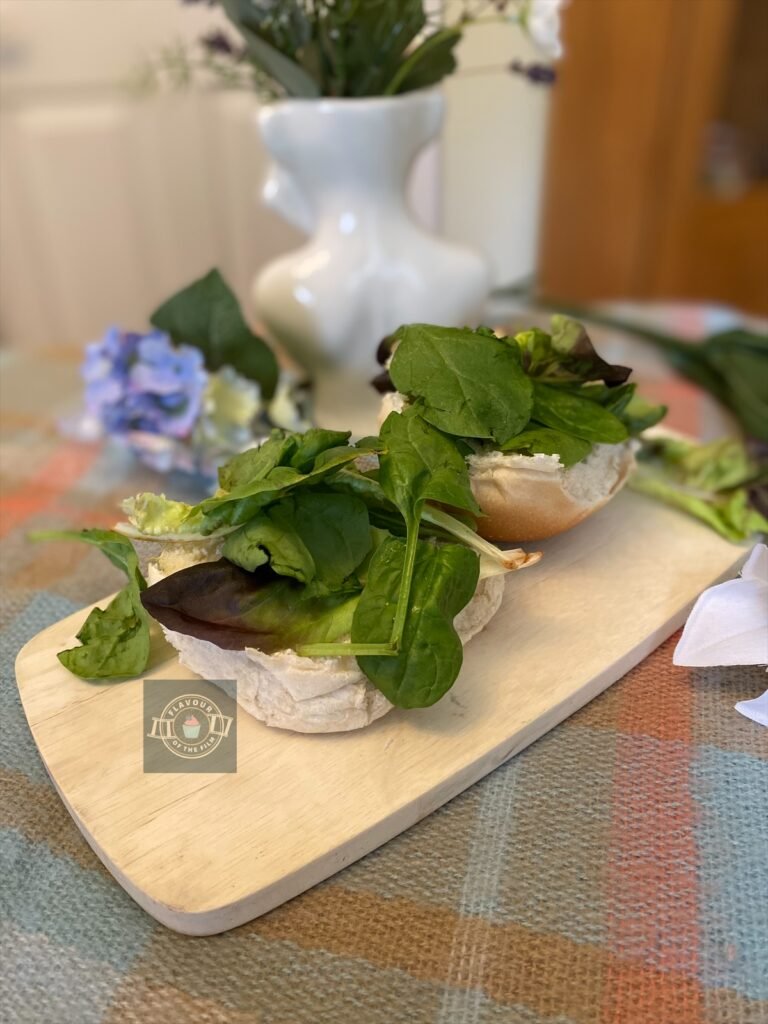 A white bread roll halved and topped with leafy greens like spinach etc, displayed on a wooden board with wildflowers around.