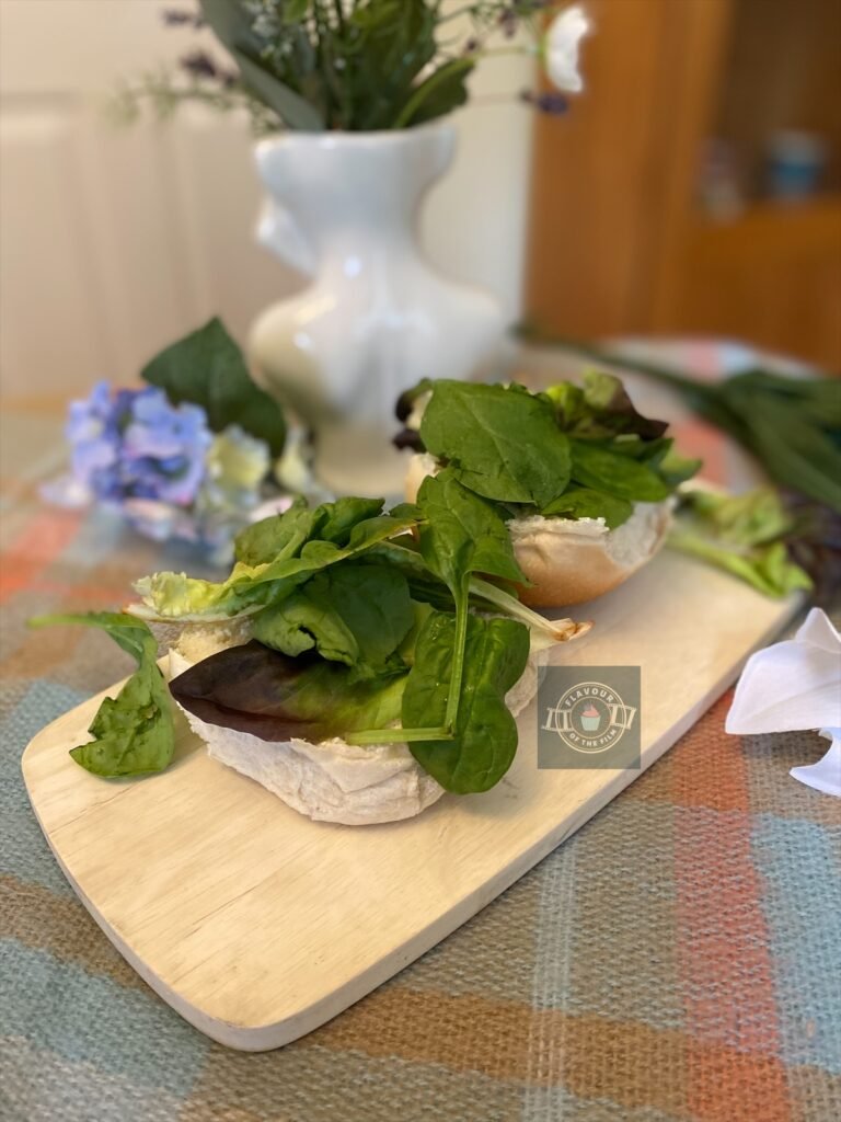 A white bread roll halved and topped with leafy greens like spinach etc, displayed on a wooden board with wildflowers around.