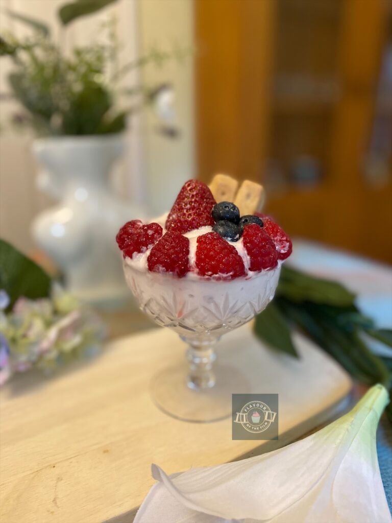 Strawberry parfait in a crystal sundae glass with fresh summer berries and shortbread pieces, displayed on a wooden board with wildflowers around.