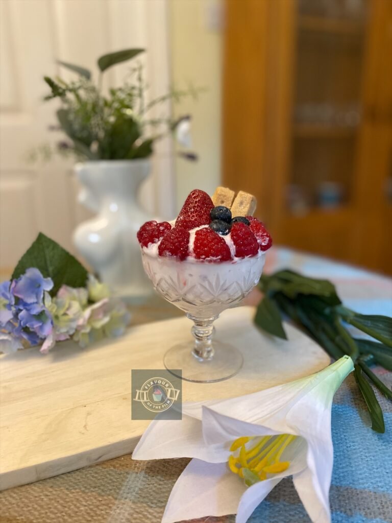 Strawberry parfait in a crystal sundae glass with fresh summer berries and shortbread pieces, displayed on a wooden board with wildflowers around.