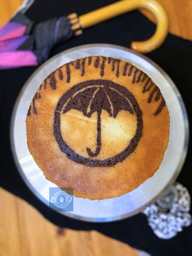 All images are of a round two layer vanilla cake with a chocolate cake design baked as part of the sponge. The design is a traced image of the umbrella tattoo from the show, with rain falling from the top. The cake is displayed on a silver cake stand. Props include a Yin Yang scrunchie, a silver wrist watch and a wooden umbrella.