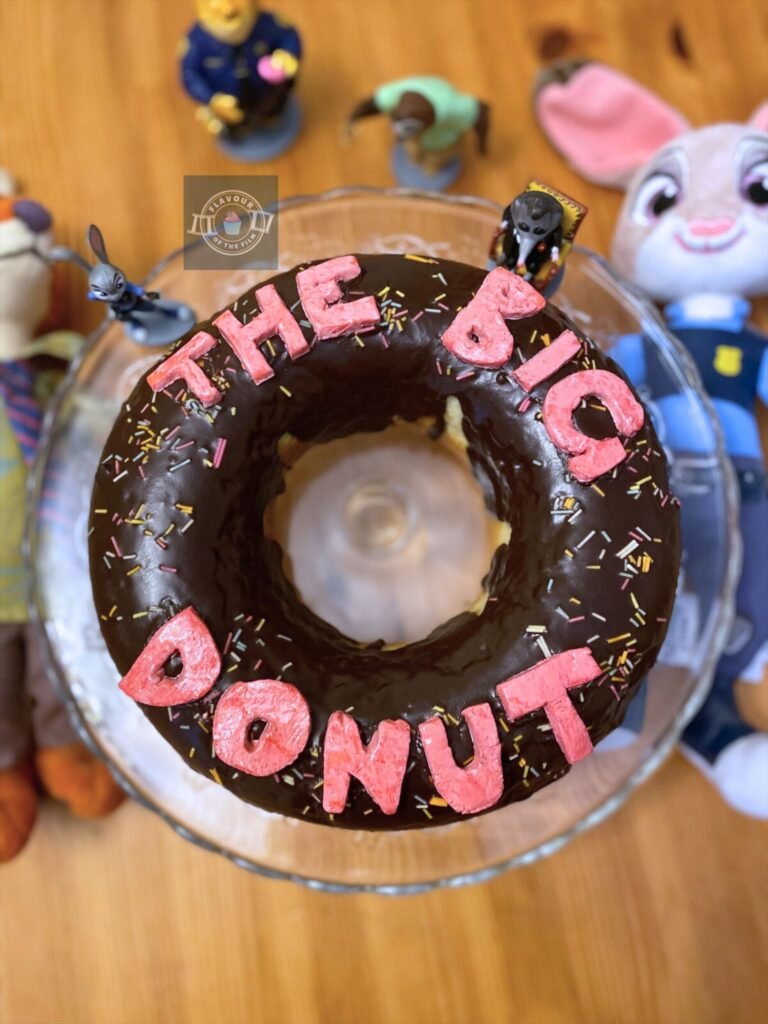 All images are of a vanilla donut shaped cake with chocolate glaze, rainbow sprinkles and "The Big Donut" on in pink fondant lettering. The cake is displayed on a glass stand. Props include Judy and Nick plushies and Zootopia character figurines.